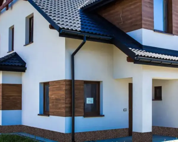 Nowoczesny dom z białym tynkiem i drewnianymi akcentami. Z czego zrobić cokół wokół domu? Widać tu kamyczki, które mogą być inspiracją.