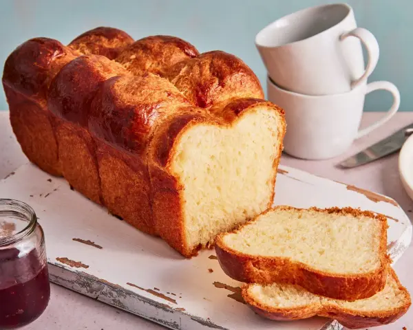 Ein goldbraunes Brioche Brot Rezept, aufgeschnitten auf einem Holzbrett mit Marmelade und Tassen im Hintergrund.