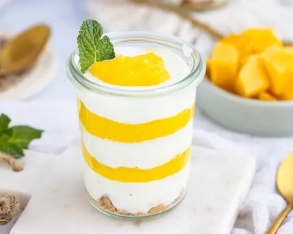 Schichtweise geschichtetes Mango-Parfait in einem Glas, garniert mit Minze. Daneben eine Schüssel mit frischen Mangostücken.