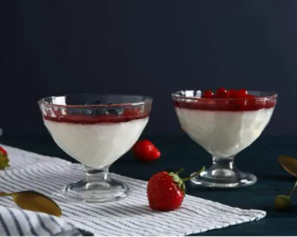 Dwie panna cotta: jedna na śmietance z borówkami, druga na jogurcie z malinami. Obok świeże truskawki i złote łyżeczki.