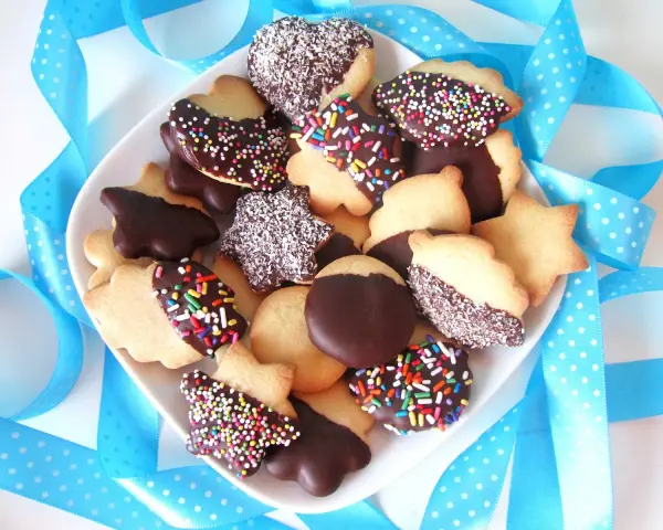 Un plato lleno de pastas de té caseras, algunas cubiertas de chocolate y chispas de colores, otras con coco rallado, rodeadas de cintas azules.