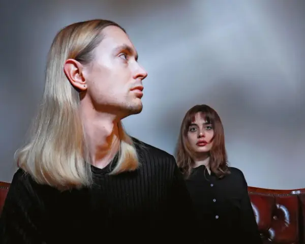 Łukasz Rozmysłowski z długimi blond włosami i kobieta z brązowymi włosami siedzą na skórzanej kanapie.