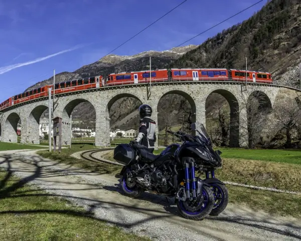 Motociclista e moto su strada sterrata, con un treno rosso che attraversa un viadotto ad archi in pietra. Paesaggio alpino con montagne sullo sfondo, vicino al passo della Novena.