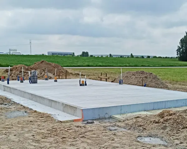 Gotowa płyta fundamentowa na budowie, otoczona piaskiem i zielenią. W tle widać pola i budynki przemysłowe.
