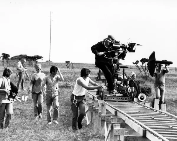 Sul set di un film, un operatore manovra una cinepresa su binari per un piano sequenza. Altri membri della troupe osservano e lavorano.