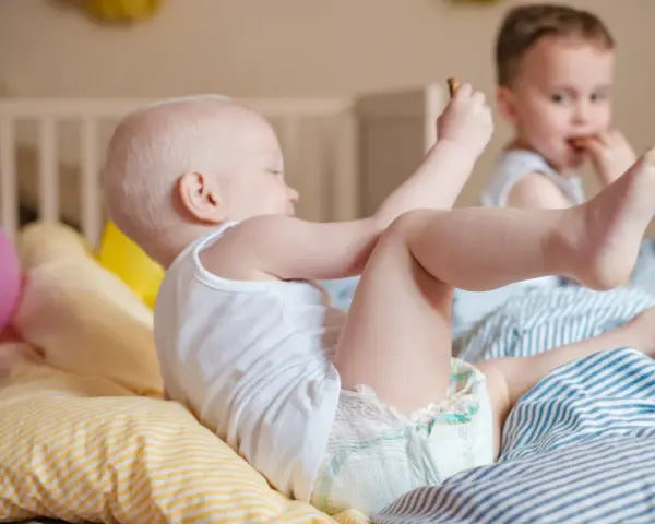 Dzieci bawią się na łóżku. Maluch w pieluszce sięga po zabawkę, a starszy chłopiec je. Ciekawe, ile pieluch miesięcznie zużywają takie energiczne maluchy!