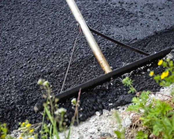 Verktyg sprider ny asfalt för att asfaltera uppfart. Grönska och gula blommor växer vid sidan.