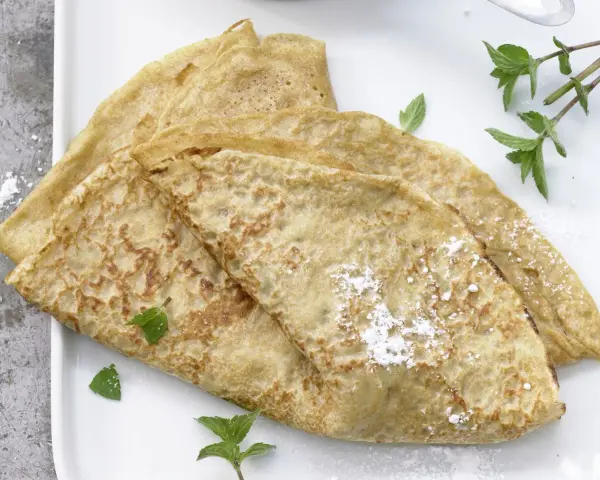 Drei goldbraune Dinkel Pfannkuchen, leicht mit Puderzucker bestäubt und mit frischer Minze garniert, auf einem weißen Teller.