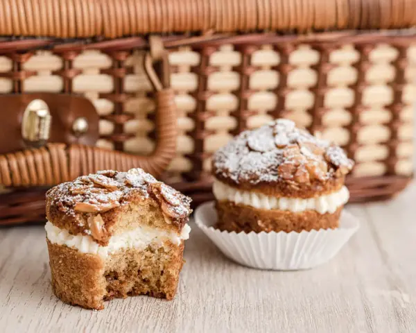Zwei leckere bienenstich muffins mit Mandel-Topping und cremiger Füllung stehen vor einem Picknickkorb.