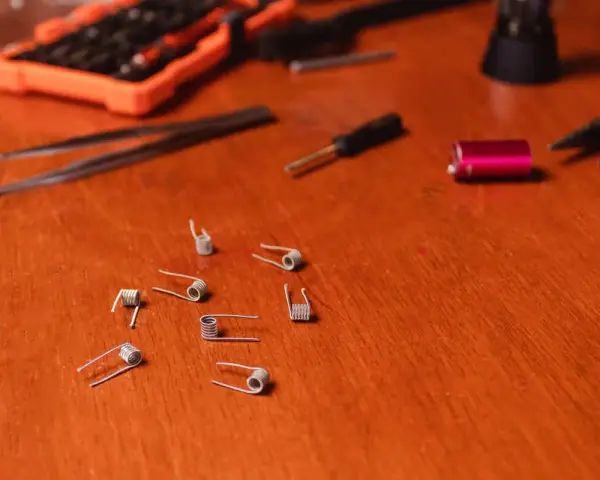 Varias resistencias comerciales de vapeo esparcidas sobre una mesa de madera, junto a herramientas como pinzas y un destornillador.