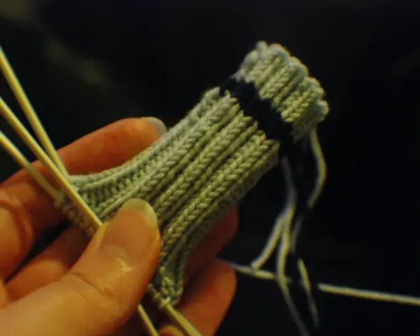 En hand håller stickor och ett ljusgrönt stickat tyg med en mörk rand. Det ser ut som att någon ska sticka avig maska.