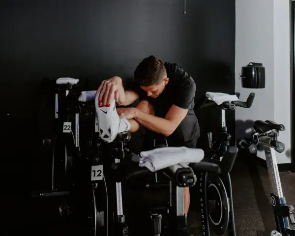 Man in black shirt stretches his outer hamstring by holding his foot, surrounded by stationary bikes in a gym.
