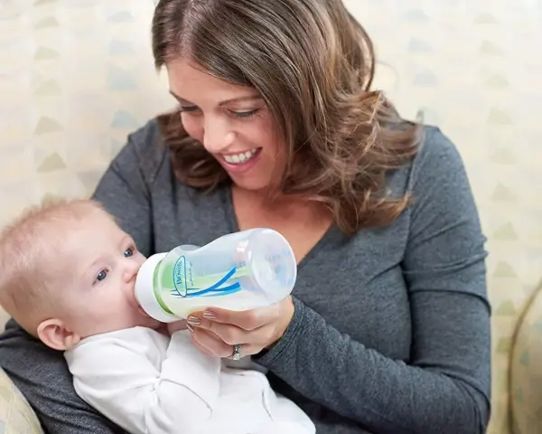 Uśmiechnięta mama karmi niemowlę butelką z systemem odpowietrzania. Smoczek do butelki Dr Brown zapewnia komfortowe karmienie.