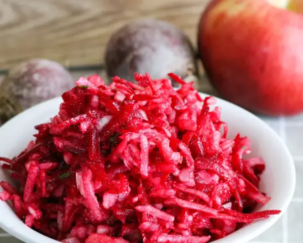 Frischer Salat aus rote Bete raspeln, mit einem Apfel und ganzen Rote Beten im Hintergrund.