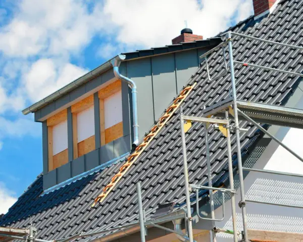 Bygga takkupa pågår. Ett hus med takpannor och byggställning, en ny takkupa med fönsteröppningar syns.