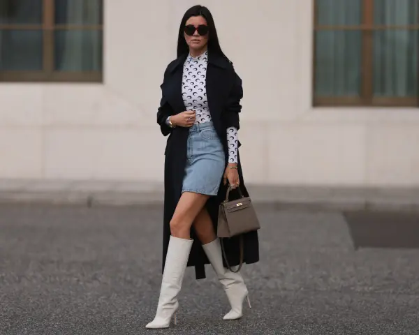 Outfit per uscire con un ragazzo: gonna di jeans, top a collo alto con stampa luna, trench nero, stivali bianchi alti e borsa grigia.