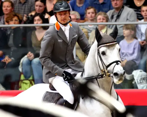 Un cavalier en tenue grise monte un cheval blanc, prêt pour la compétition. Le public est attentif.
