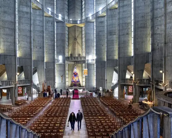 Intérieur de la cathédrale de Royan, avec ses murs en béton brut, ses rangées de chaises et son vitrail coloré au fond.