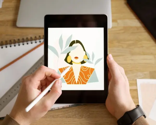 Ręce trzymają tablet z rysunkiem kobiety, tworzonym w aplikacji procreate. Obok leżą szkicownik i ołówek.