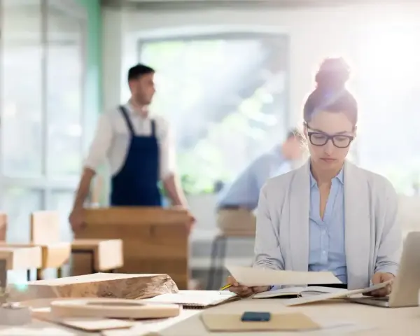 Kobieta w okularach analizuje dokumenty przy biurku, podczas gdy w tle mężczyzna w fartuchu pracuje przy drewnie. To jej staż z urzędu pracy.