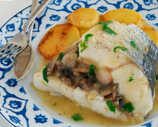 Deliciosa merluza con gambas y champiñones, servida con patatas doradas en un plato azul y blanco.