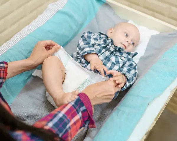 Mama sprawdza, kiedy zmienić rozmiar pieluchy maluszkowi. Dziecko leży na przewijaku w kratkę.
