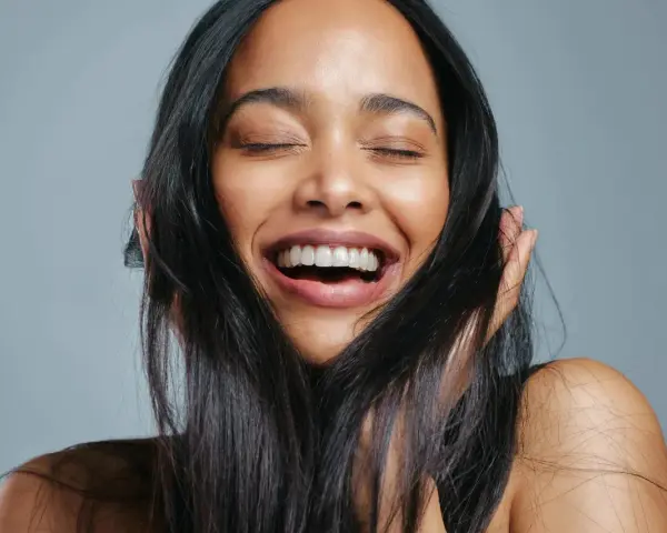 Une femme rit aux éclats, ses yeux fermés. Ses cheveux noirs encadrent son visage radieux, témoignant de la beauté que la vitamine B7 peut apporter.