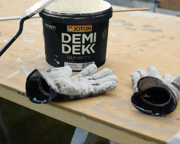 Förbereder för att måla utomhus på våren. En burk Jotun Demi Dekk, pensel och slitna handskar ligger på ett träbord.