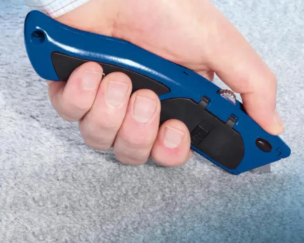 Hand hält blauen Teppichmesser, um grauen Teppich zu schneiden. Präzises Arbeiten beim Teppich verlegen.