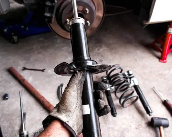 Mano guantata tiene un ammortizzatore nuovo, pronto per sostituire quelli scarichi dell'auto. Attrezzi e molle a vista.
