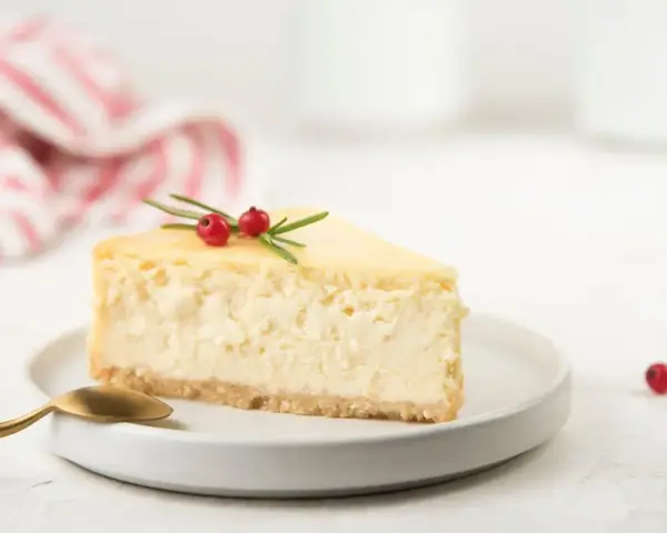 Una fetta di cheesecake con yogurt greco, guarnita con rosmarino e bacche rosse, su un piatto bianco con cucchiaino dorato.