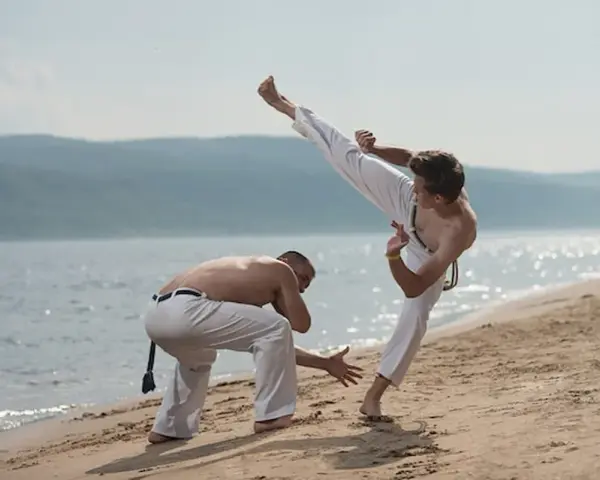 Dwóch mężczyzn ćwiczy capoeirę na plaży. Jeden wykonuje wysokie kopnięcie, drugi jest w pozycji obronnej. Widać ich płynny ginga styl gry.