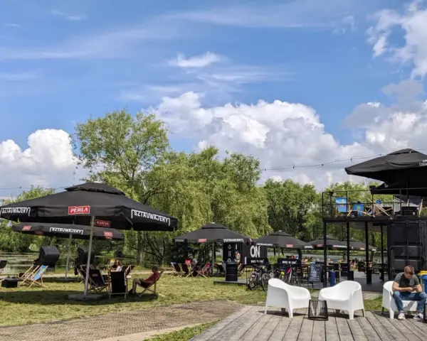 Relaks nad wodą w **Dolinie Trzech Stawów**. Ludzie odpoczywają pod parasolami, a w tle widać zielone drzewa i błękitne niebo z chmurami.