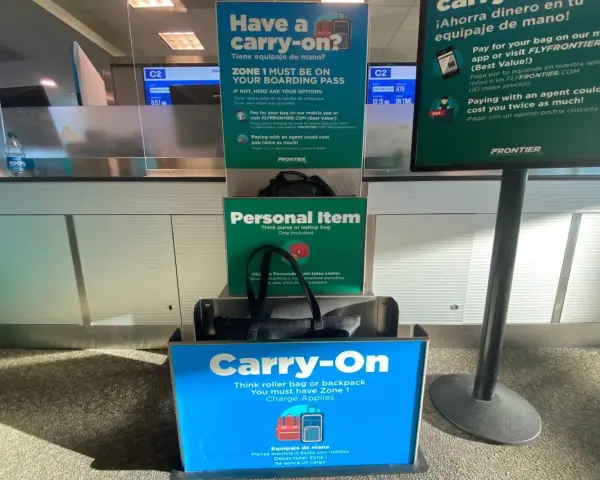 Frontier Airlines personal item size guide at the airport, showing a tote bag fitting in the "Personal Item" box and a roller bag in the "Carry-On" box.