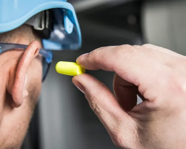 Main tenant, casque bleu, lunettes de protection et bouchon d'oreille jaune pour se protéger d'un bruit de 120 décibels équivalent.