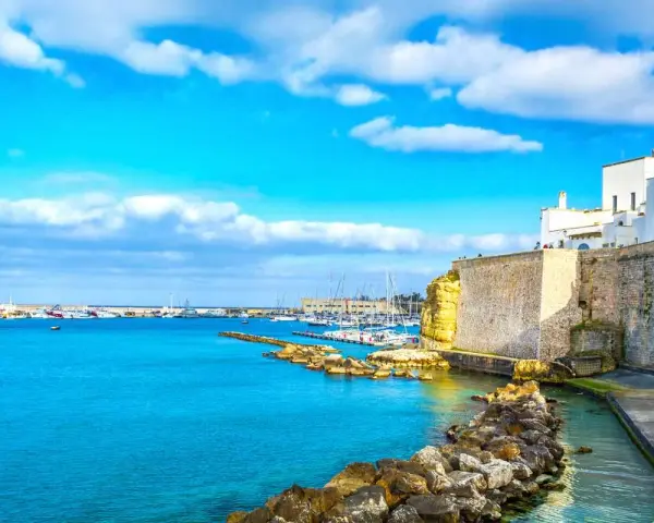 Un incantevole cammino del Salento: un porto con barche a vela, case bianche su scogliere dorate e un mare azzurro sotto un cielo limpido.