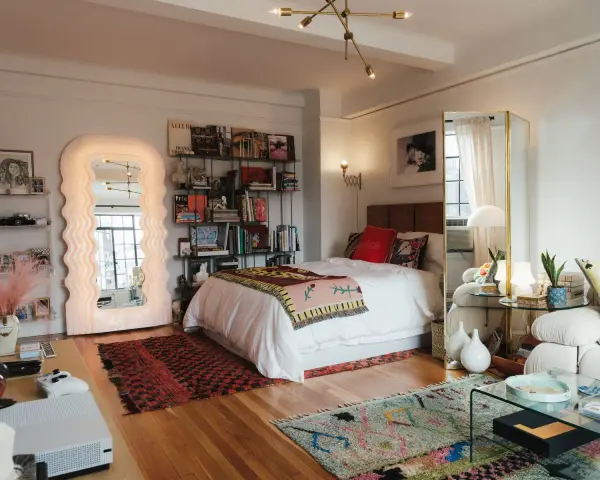 Ideas para decorar habitación estudio: cama blanca, espejo ondulado, estanterías con libros y arte, alfombras coloridas y un sofá acogedor.