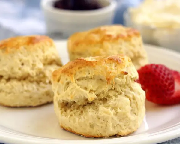 Tre gyllene engelska scones på en vit tallrik med en jordgubbe, sylt och grädde i bakgrunden.