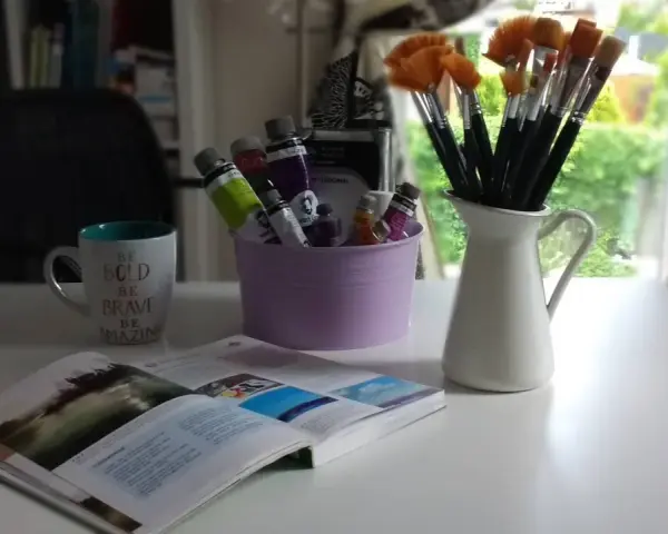 Pędzelki malarskie w dzbanku, tubki farb w fioletowym wiaderku, kubek z napisem i otwarta książka.
