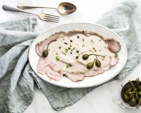 Eleganckie vitello tonnato z kremowym sosem, kaparami i natką pietruszki na białym talerzu, obok srebrnych sztućców i lnianej serwetki.