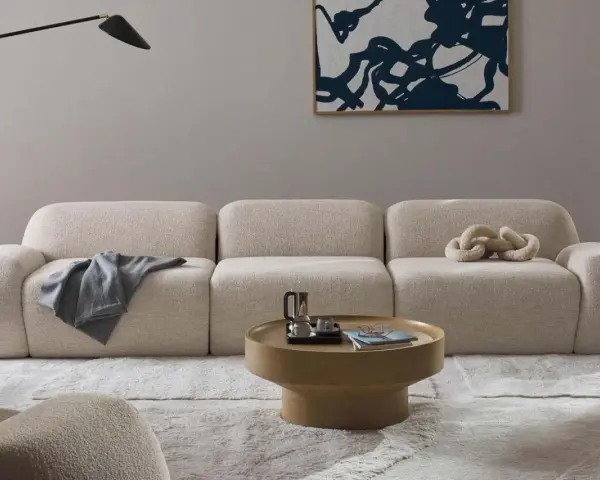 Divano modulare in tessuto bouclé color crema, con cuscino decorativo a forma di catena. Il materiale del divano è morbido e invitante, perfetto per un salotto moderno.