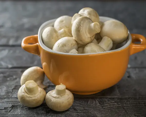 Pomarańczowa miseczka pełna świeżych pieczarek, jakby w ciąży z nowymi smakami. Kilka pieczarek leży obok na drewnianym blacie.