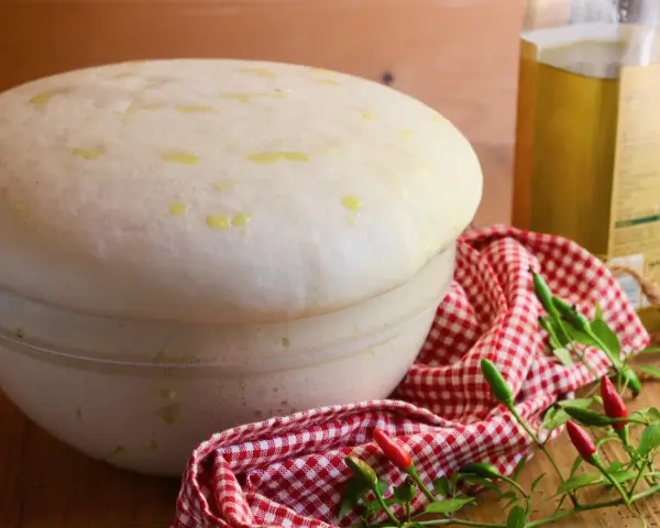 Impasto lievitato per pizza con lievito di birra, soffice e profumato, pronto per essere infornato. Olio e peperoncini completano la scena.