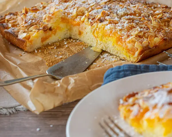 Duże ciasto z brzoskwiniami i migdałami, posypane cukrem pudrem, na papierze do pieczenia.