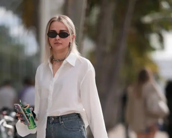 Donna con occhiali da sole, collana, camicia bianca e jeans, tiene in mano una lattina verde e un telefono.