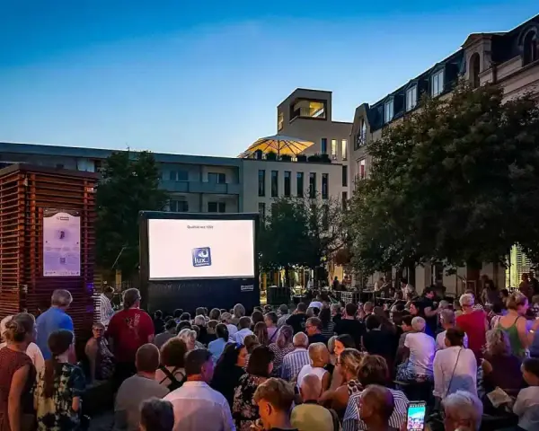 Ein lauer Sommerabend in Berlin: Menschen genießen ein Open-Air-Kino-Erlebnis vor einer Leinwand mit dem Logo "Lux Augenoptik".
