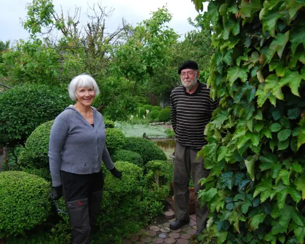 A smiling woman and a man stand in a lush garden with manicured bushes and trees.