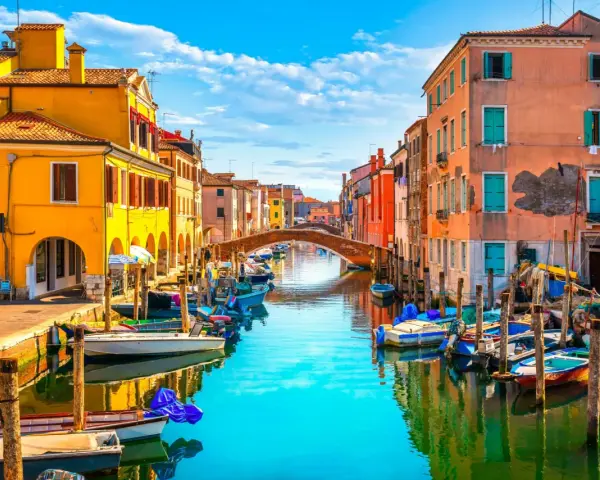 Colorate case affacciate su un canale con barche ormeggiate. Un ponte ad arco collega le due sponde. Un'immagine che ispira su cosa fare a Chioggia.