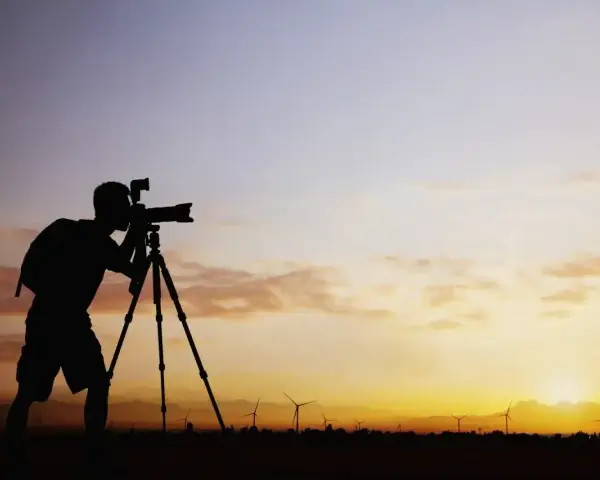 Sylwetka fotografa z aparatem na statywie na tle zachodzącego słońca i farmy wiatrowej. Zastanawiam się, ile bierze fotograf za 18 takich ujęć.