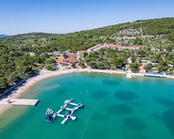 Malownicza zatoka z turkusową wodą, plażą i wodnym placem zabaw. W tle widać zabudowania i zielone wzgórza, idealne na camping Murter.